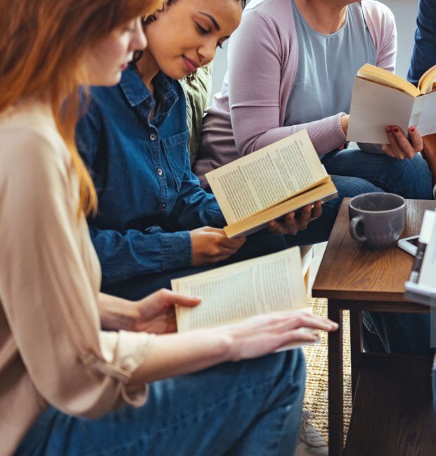 Small group of people with a mixed age range sitting at a table, discussing and reading books together.